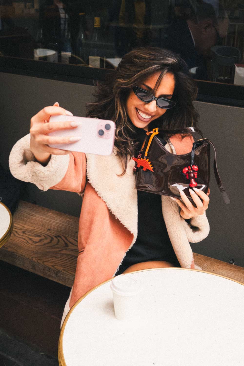 Woman taking a selfie with a phone and holding a brown handbag with a red lobster and brown dinosaur shaped handmade leather bag charm