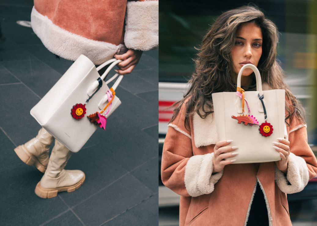 Woman holding a white handbag with colorful charms, wearing a brown coat with white fur trim.