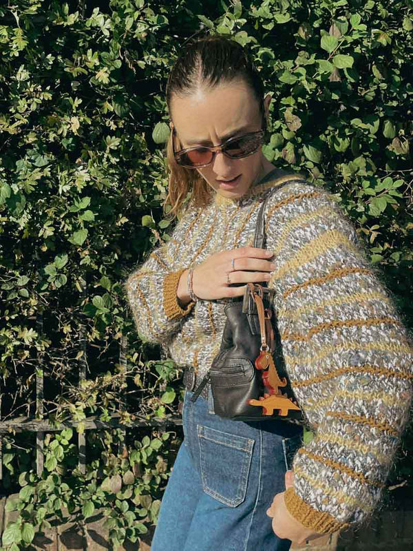 Women holding a handbag with bag charms and wearing a patterned sweater and sunglasses, standing against a green leafy background