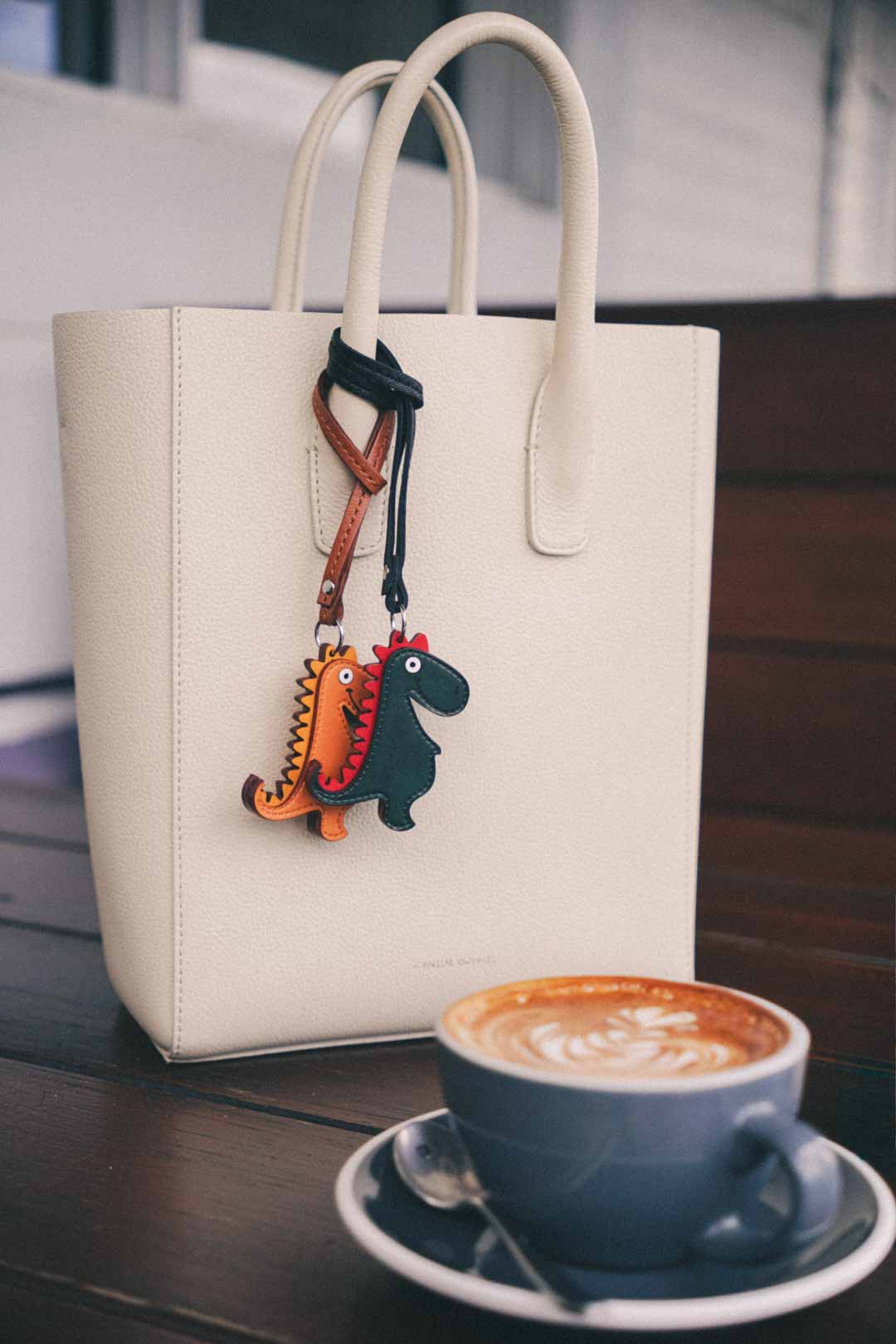 White handbag with two dinosaur shaped handmade leather bag charm next to a cup of coffee on a wooden table.