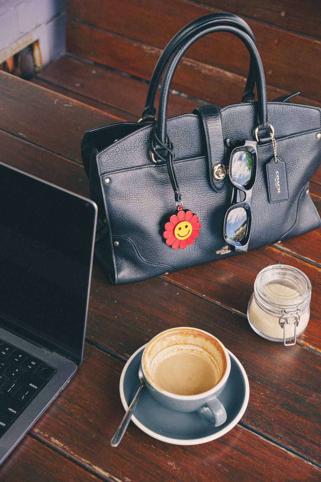 Black handbag with a smiling flower handmade leather bag charm on a wooden table with a laptop, coffee cup, and jar.