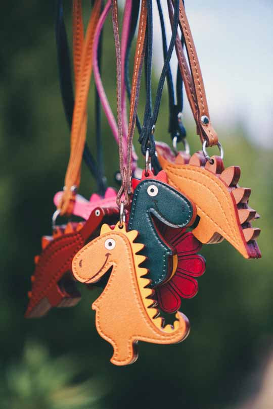 Colorful dinosaur-shaped handmade leather bag charms hanging against a blurred green background