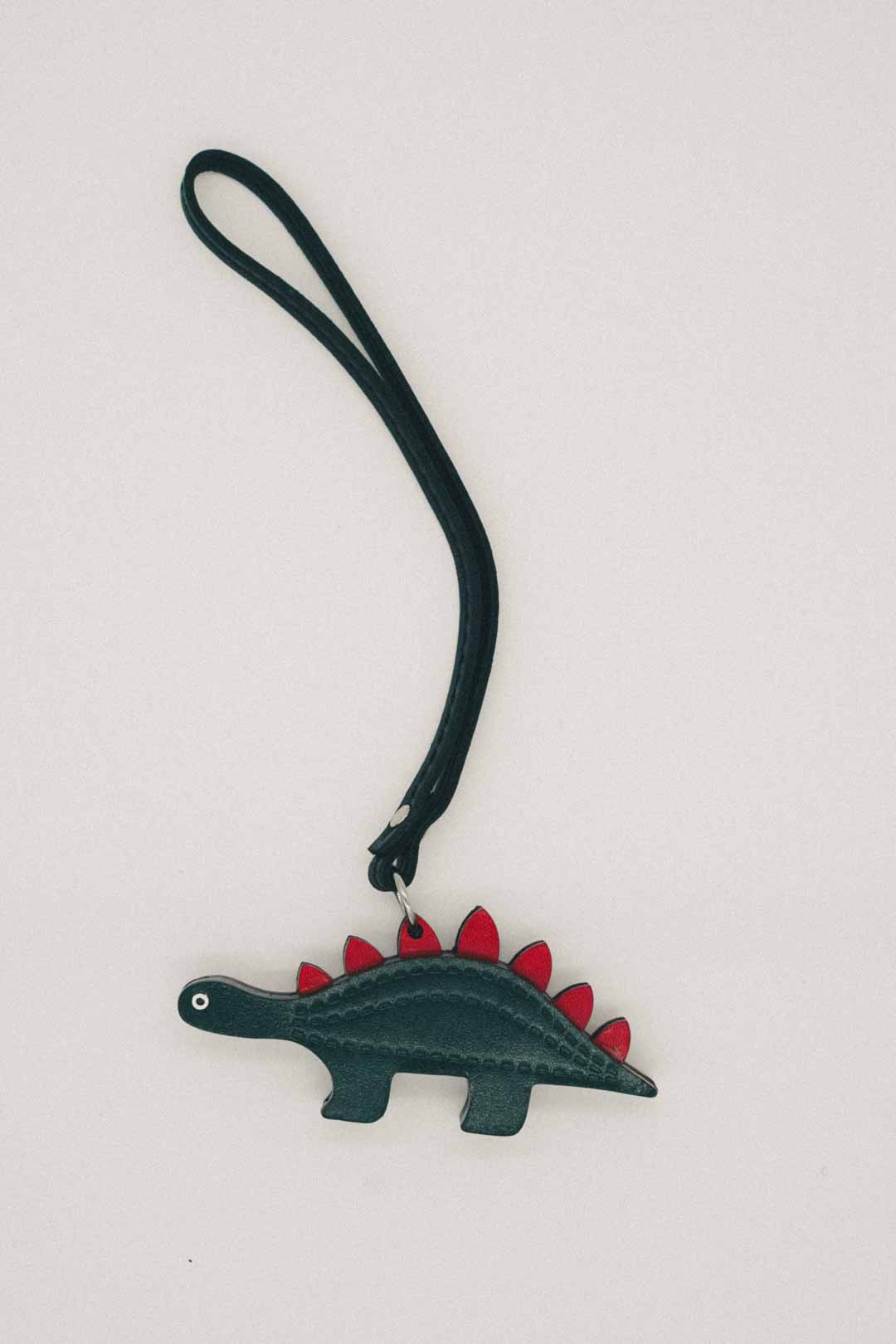 Dark green dinosaur-shaped handmade leather bag charm with red spikes on a white background