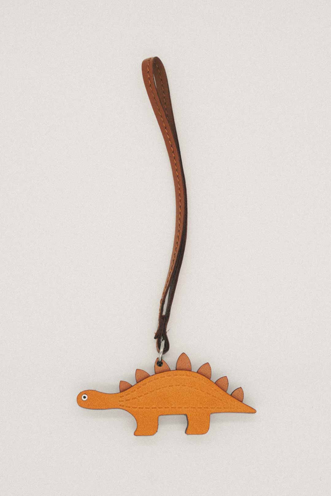 Light brown dinosaur-shaped handmade leather bag charm with a brown strap on a light gray background