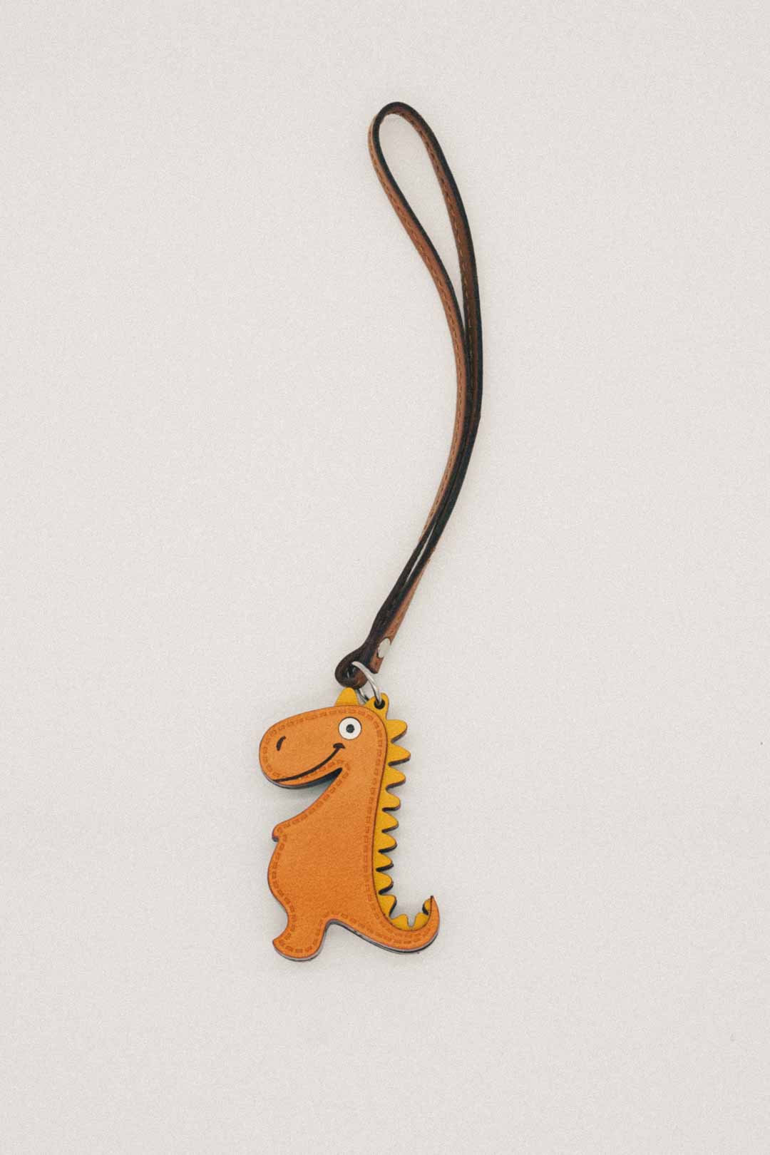Yellow dinosaur-shaped handmade leather bag charm with a brown strap on a light gray background