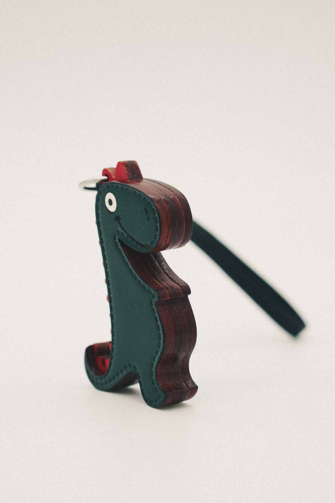 Side view of a green and brown handmade leather bag charm shaped like a dinosaur on a white background