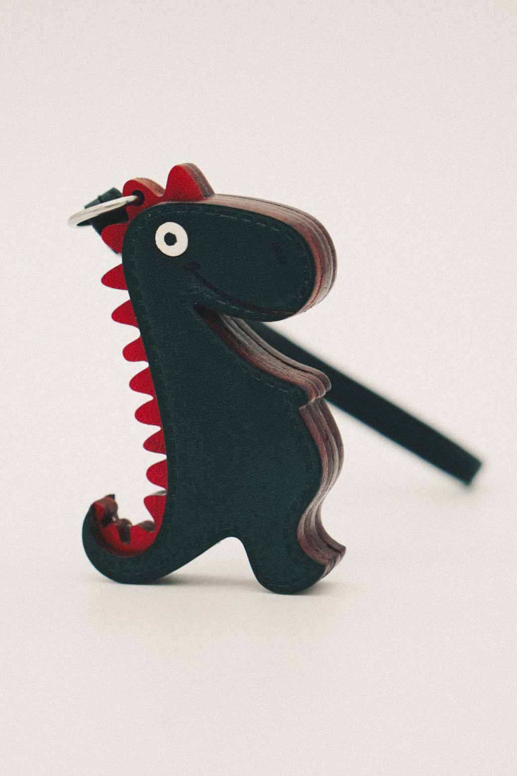 Dark green dinosaur-shaped handmade leather bag charm with red accents on a white background