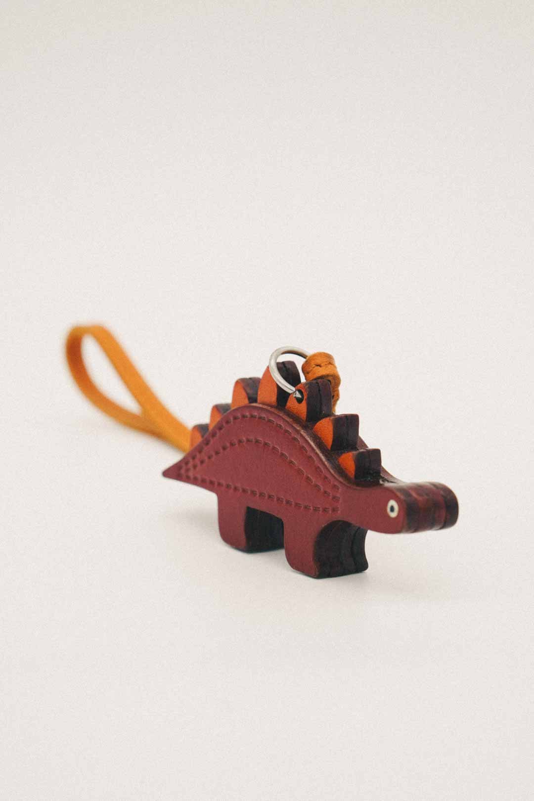 Brown dinosaur-shaped handmade leather bag charm with orange strap on a white background