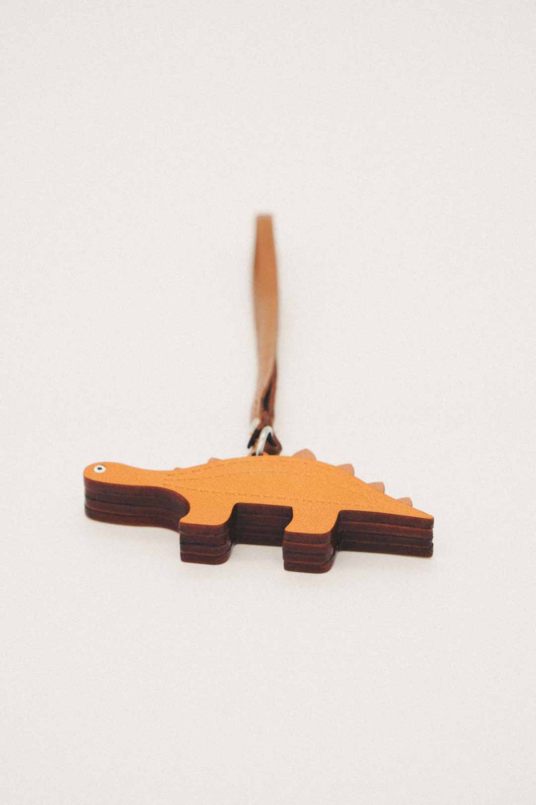 Bottom view of dinosaur-shaped handmade leather bag charm with a strap on a light gray background