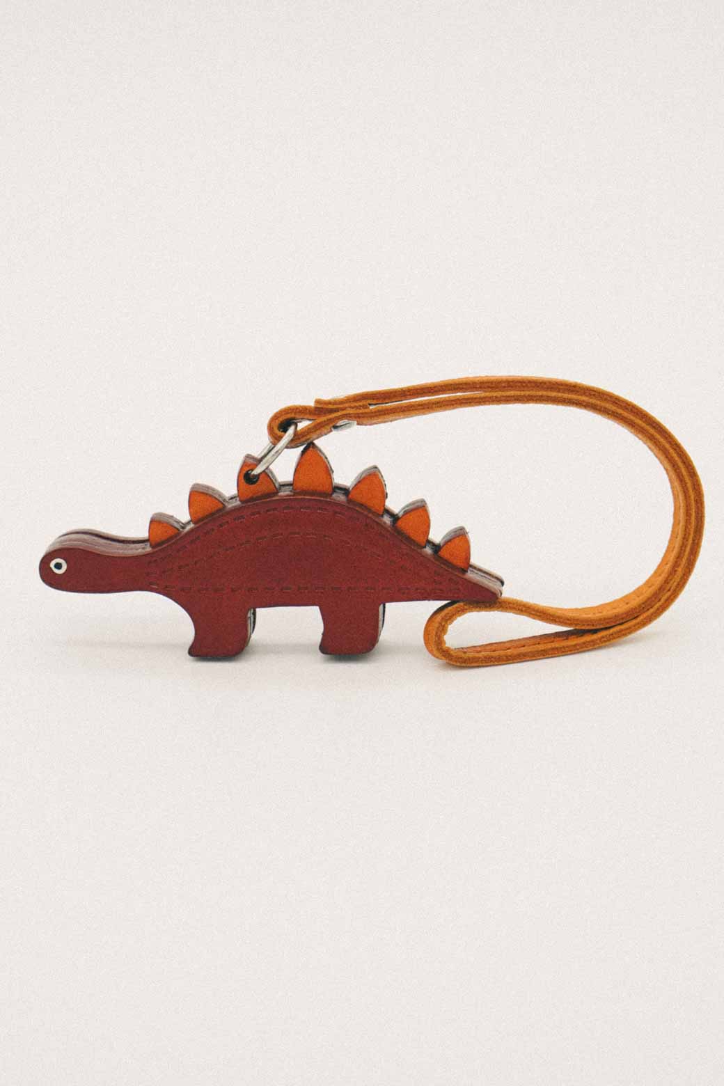 Brown dinosaur-shaped handmade leather bag charm with a brown body and orange spikes on a white background