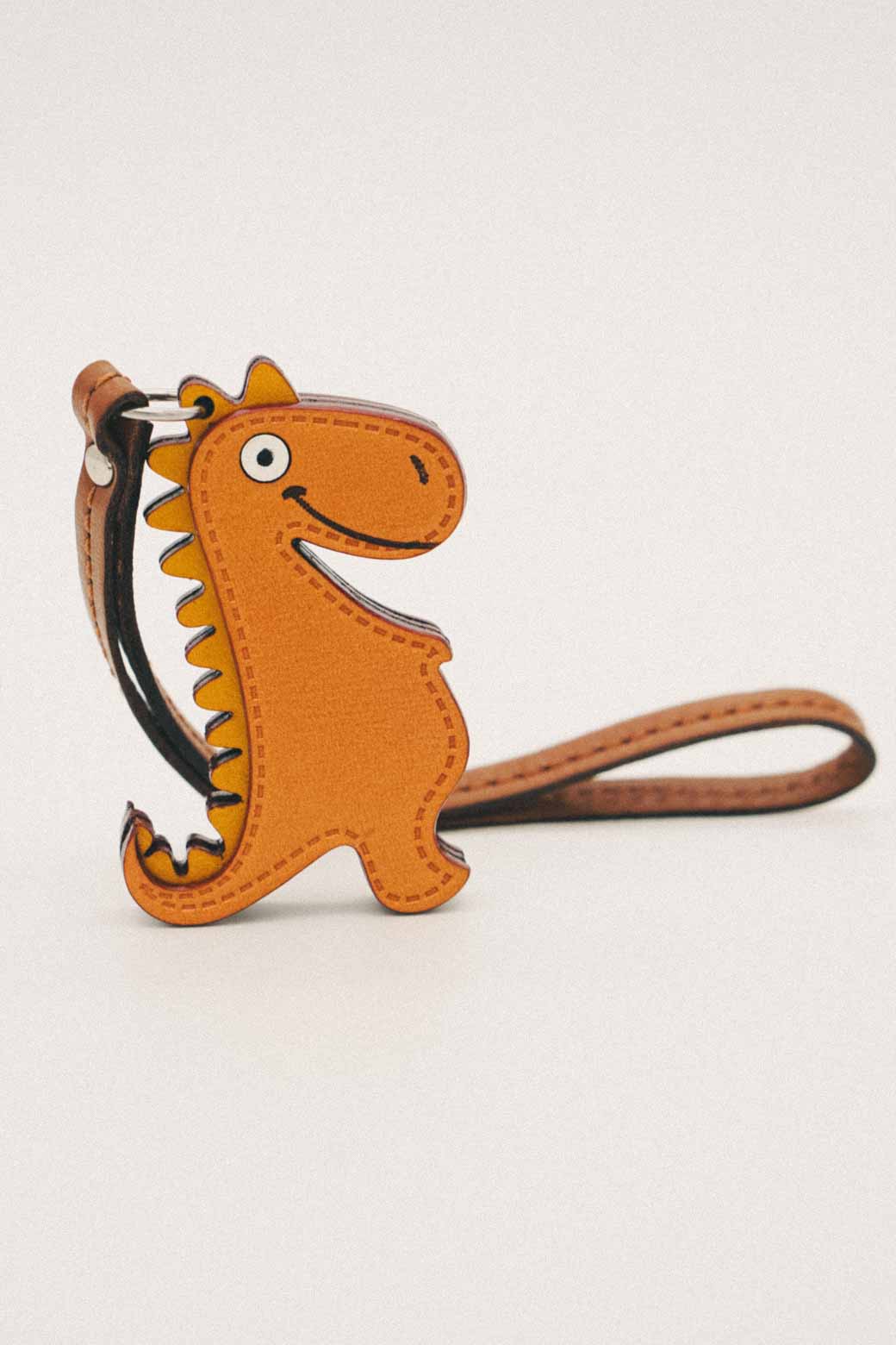 Golden orange dinosaur-shaped handmade leather bag charm with a brown strap on a light gray background