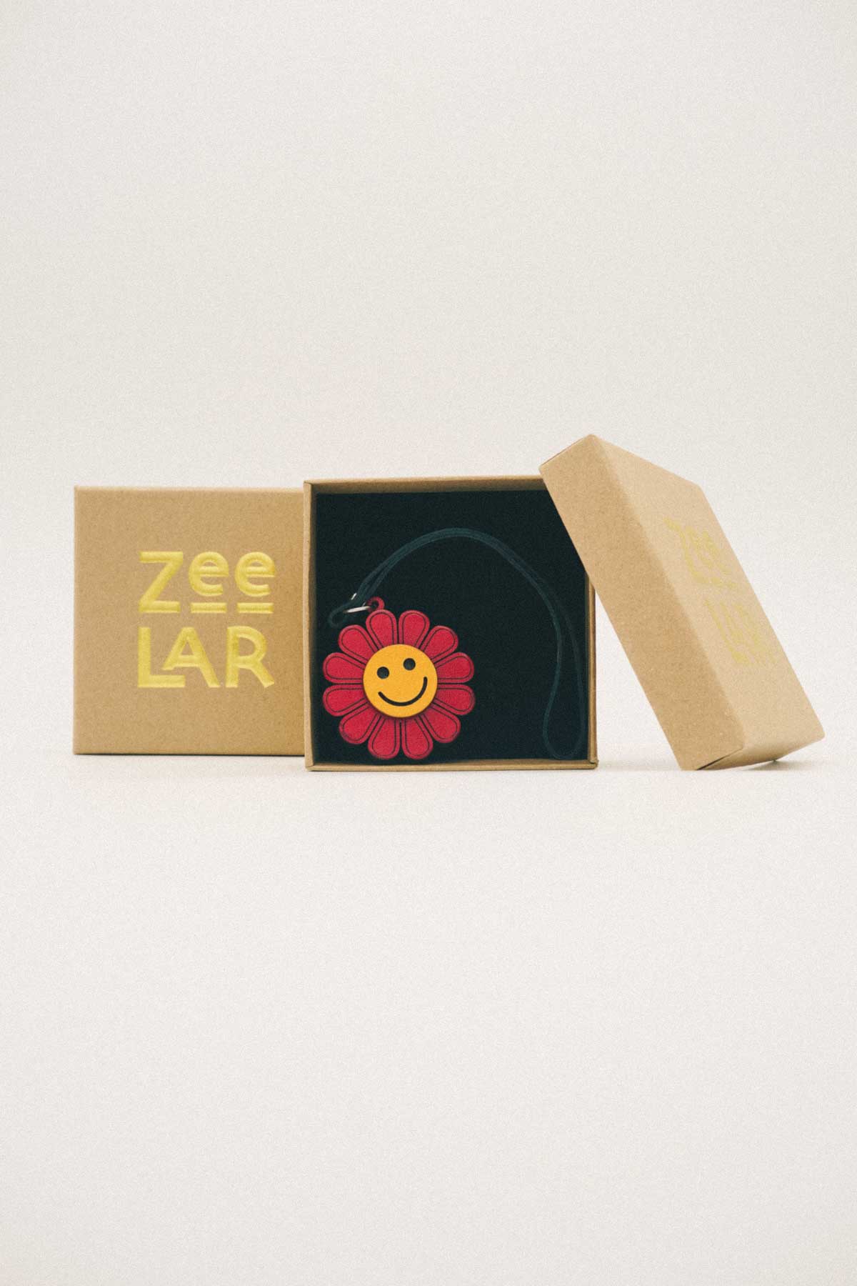 Box containing a smiley face flower handmade leather bag charm and 'Zeelar' branding on a light background