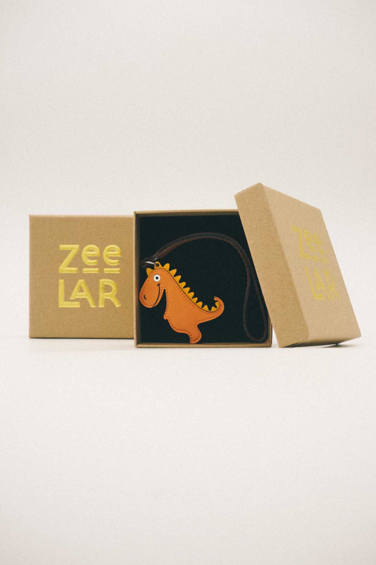 Box containing a golden orange dinosaur shaped handmade leather bag charm and 'Zeelar' branding on a white background
