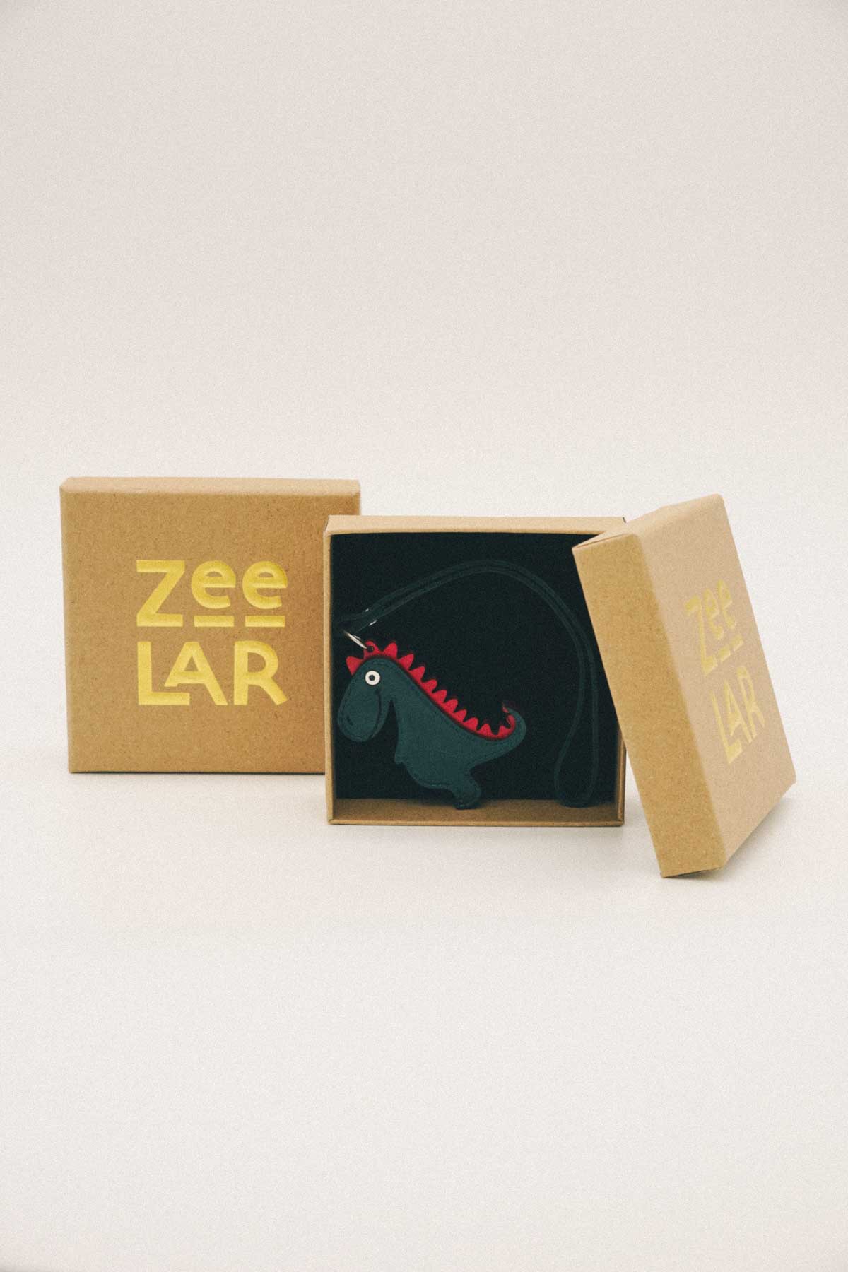 Dark green dinosaur shaped handmade leather bag charm inside a box with 'Zeelar' branding on a white background