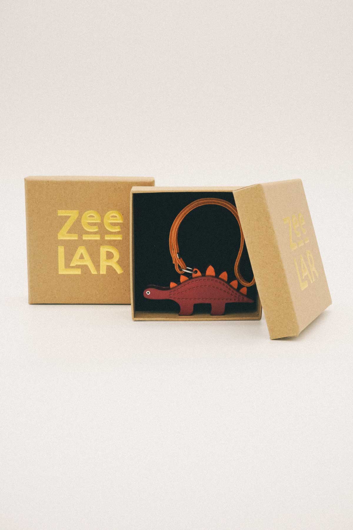 Box containing a brown handmade leather bag charm in the shape of a dinosaur with 'Zeelar' branding on a white background