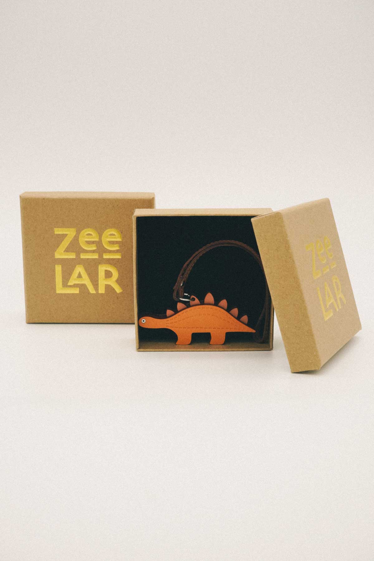 Open brown box containing orange handmade leather bag charm on a white background, featuring the brand 'Zeelar'.