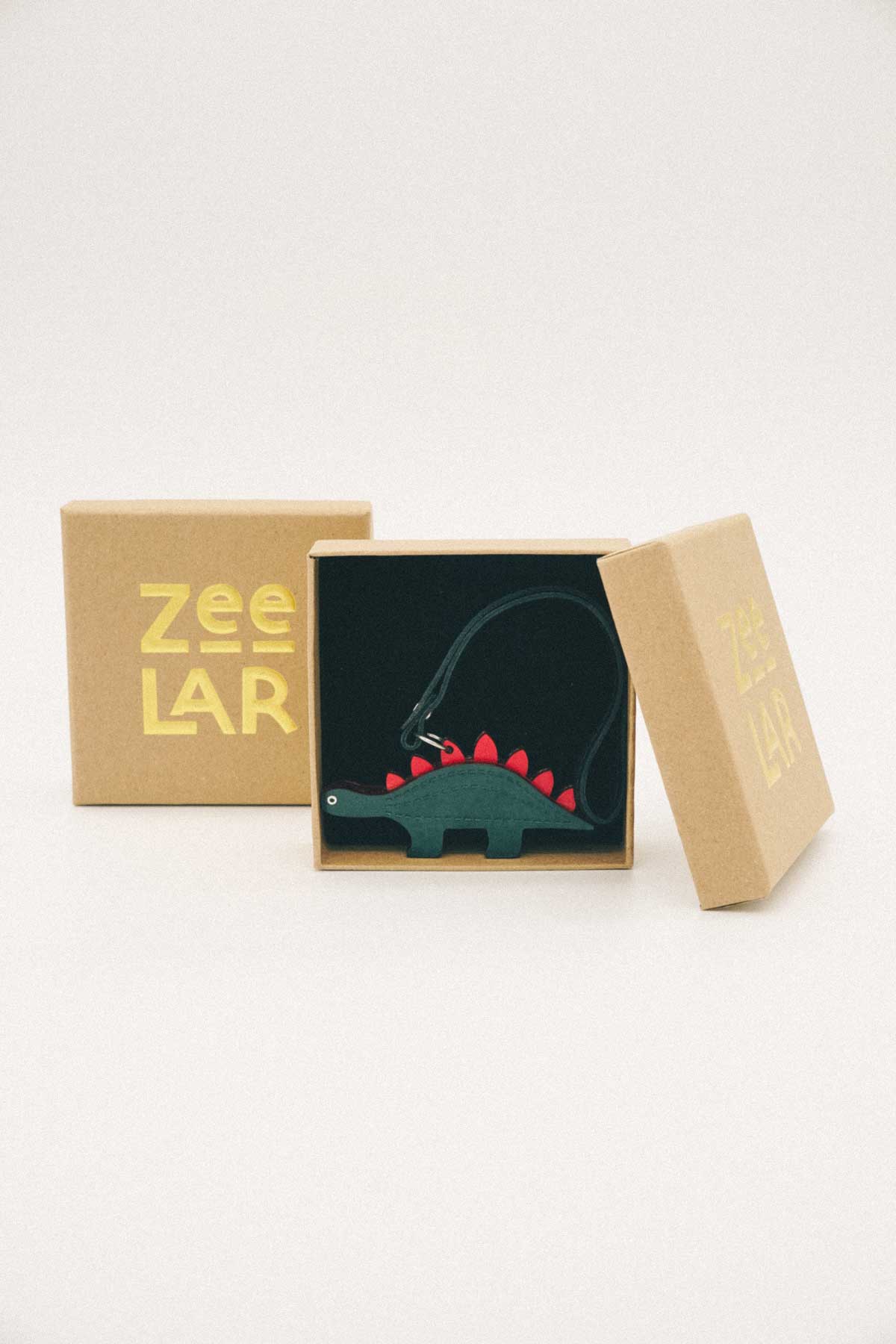 Dark green dinosaur handmade leather bag charm in a cardboard box with 'Zeelar' branding on a white background