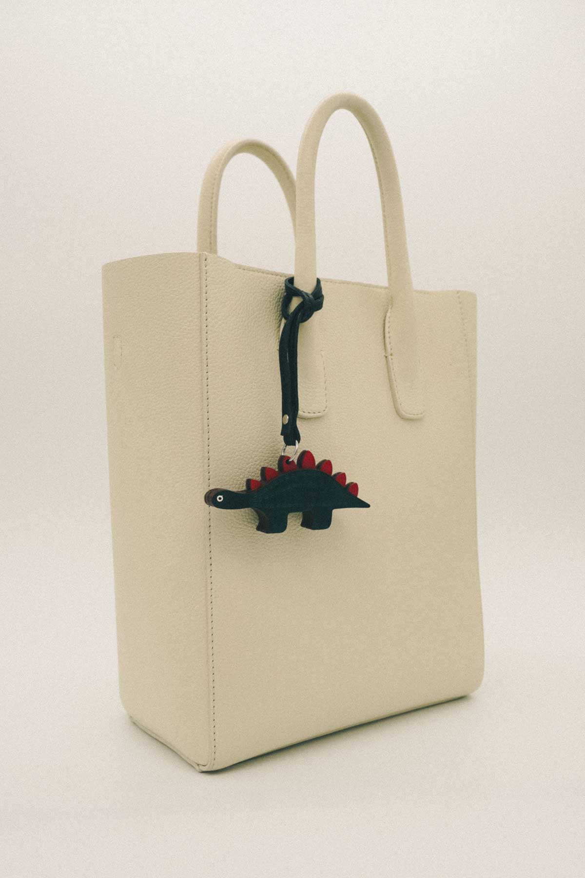 Beige tote bag with a dark green dinosaur handmade leather bag charm on a white background