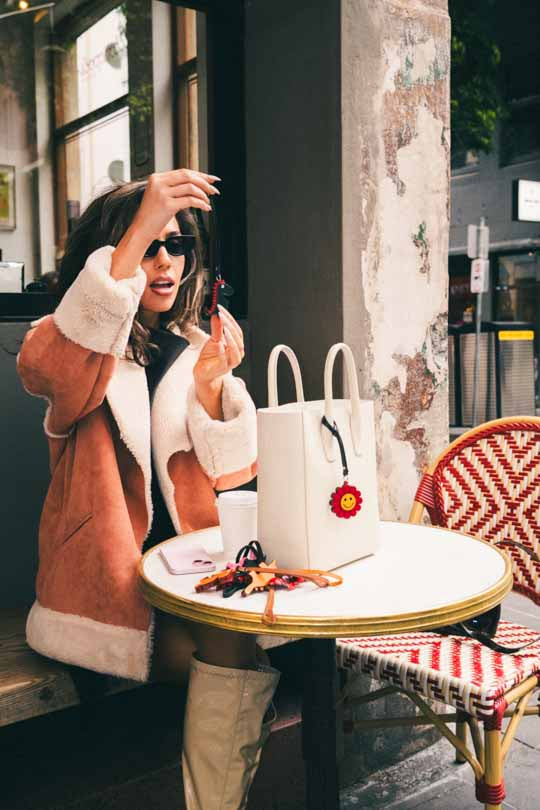 Woman sitting at a cafe table with a white bag, wearing a brown coat and sunglasses and holding a green handmade leather bag charm