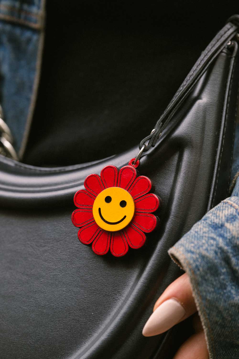 Red and yellow flower-shaped handmade leather bag charm with a smiley face on a black bag held by a person wearing blue jeans.