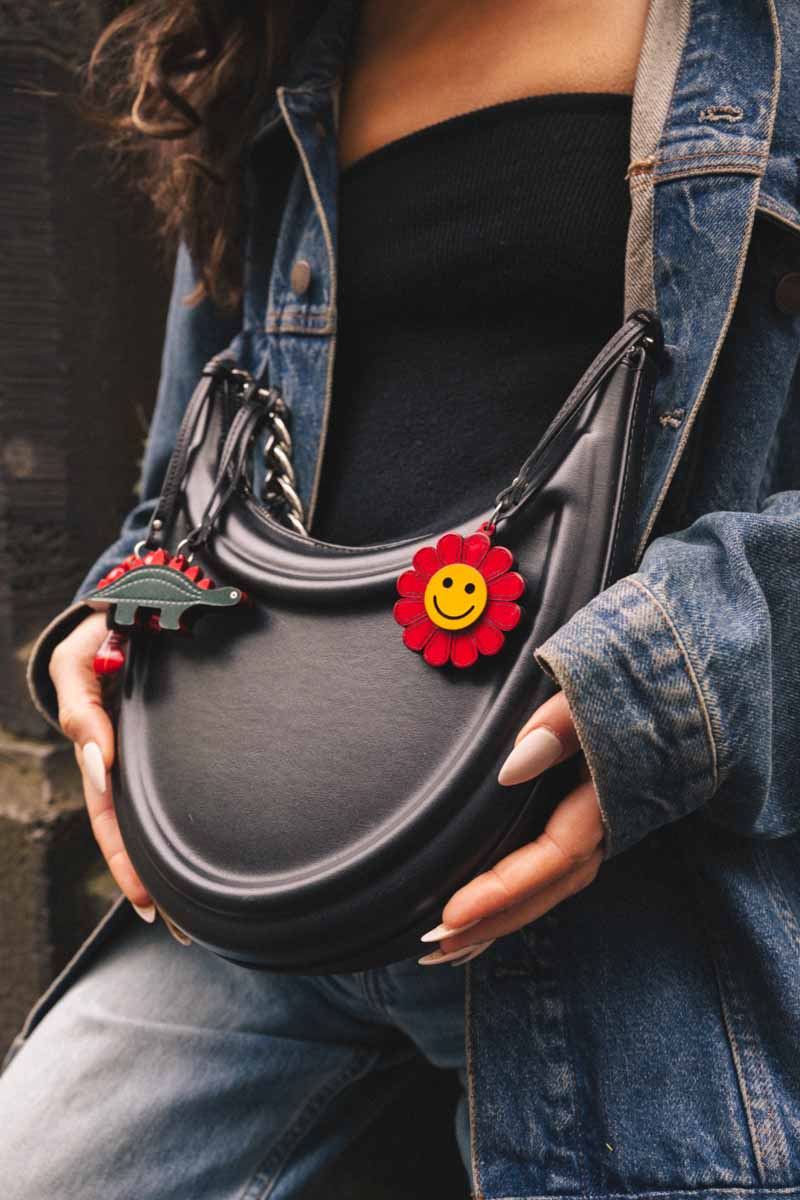 Person holding a black handbag with a red smiling flower handmade leather bag charm, wearing a denim jacket.