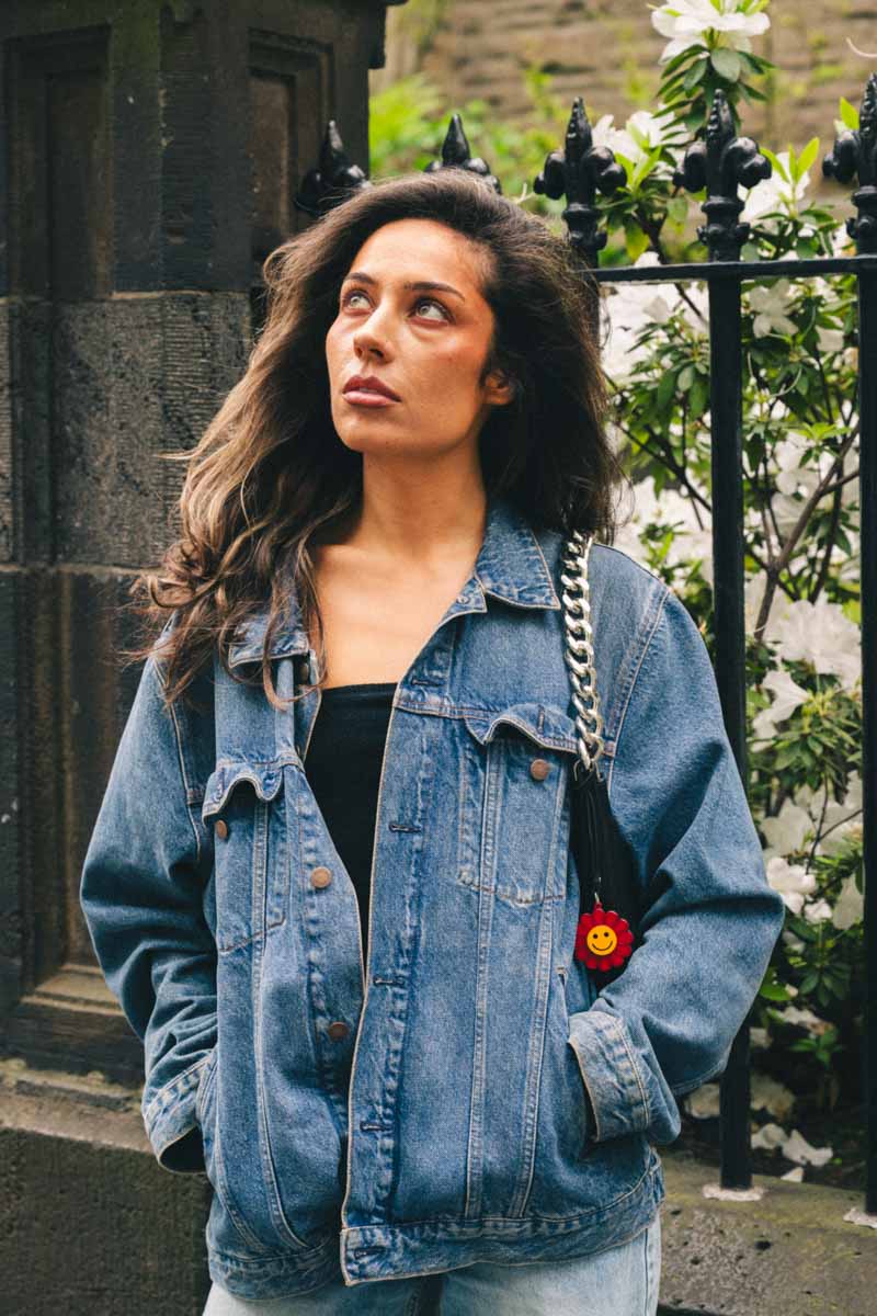 Woman wearing a denim jacket with a smiley face flower bag charm, standing outdoors.