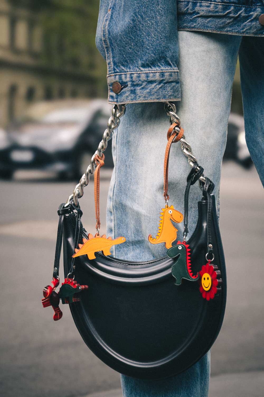 Black handbag with colorful dinosaur, lobster and flower shaped handmade leather bag charm held by a person on a street.