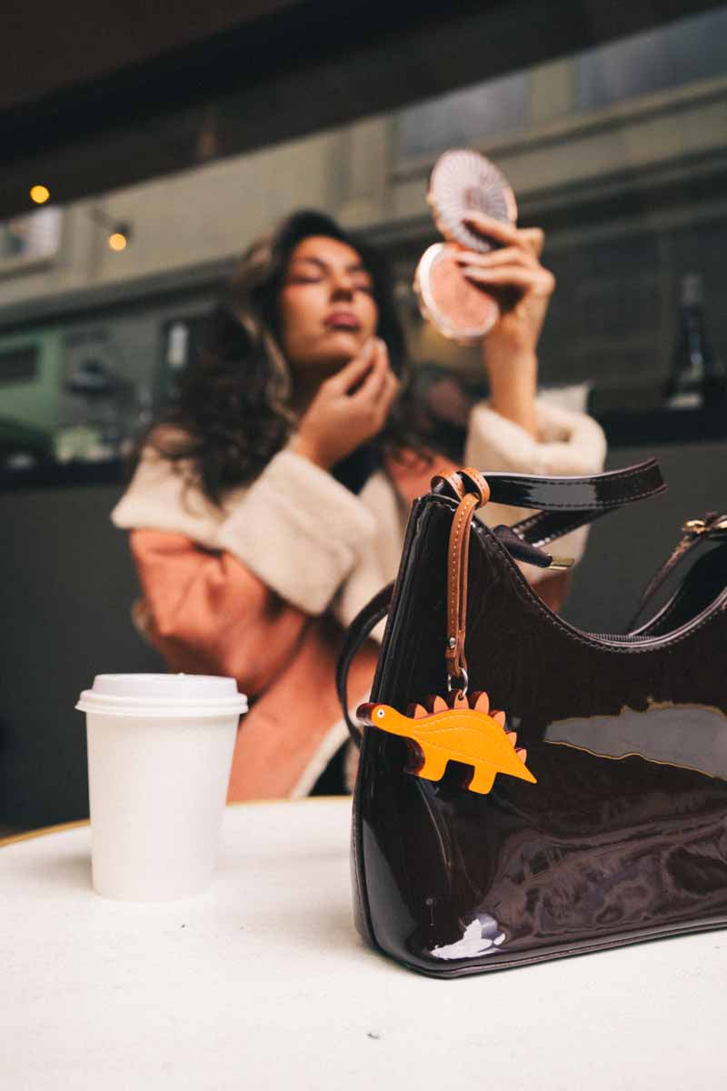 Brown bag with yellow dinosaur handmade leather bag charm on a table with woman doing makeup in background