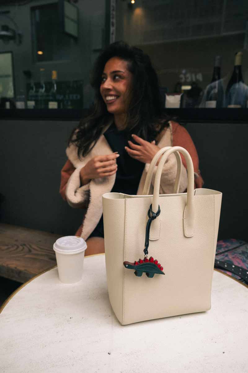 Woman sitting at a table with a beige tote bag featuring a dinosaur handmade leather bag charm