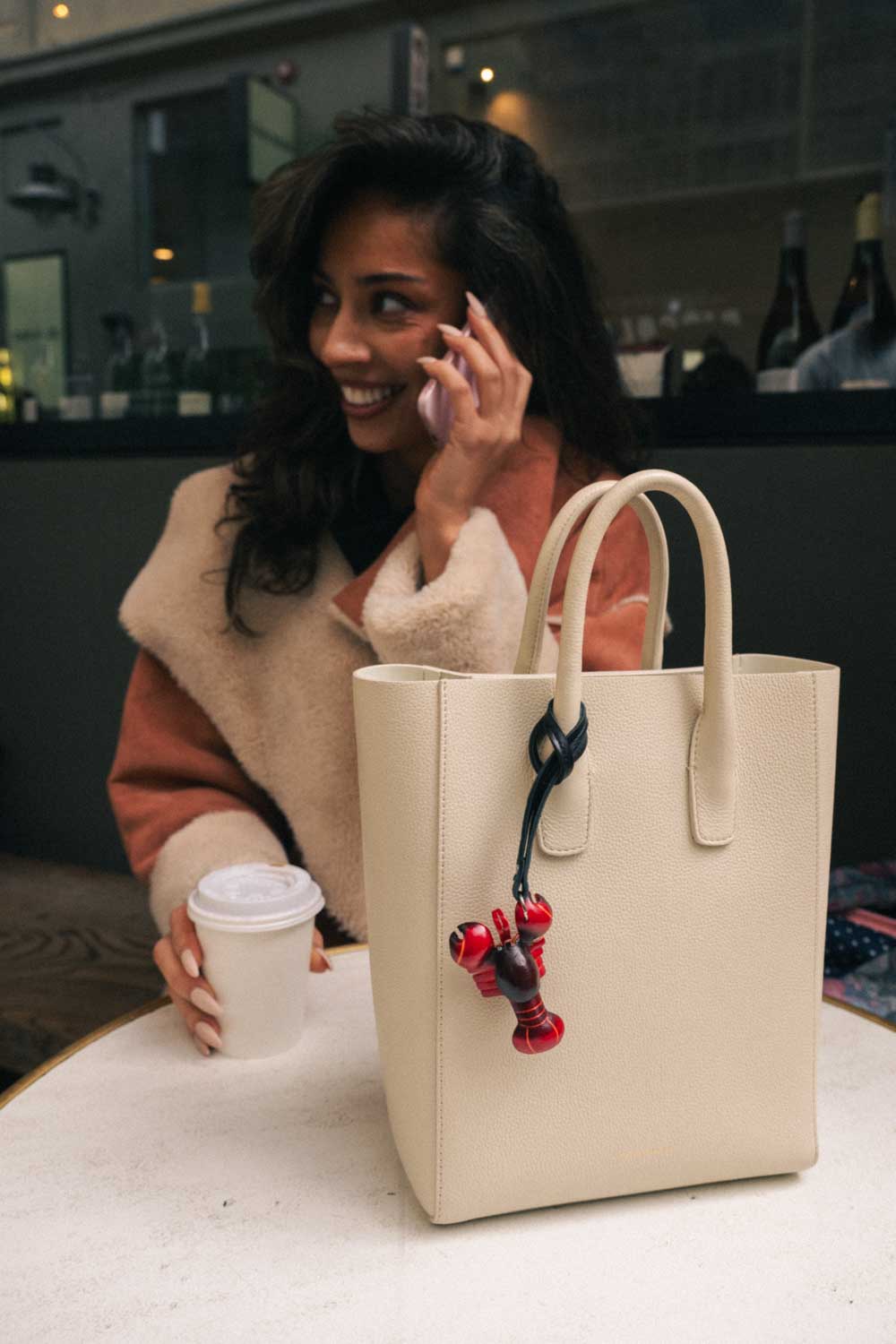 Woman talking on phone with a beige handbag featuring a red and burgundy lobster handmade leather bag charm on a table.