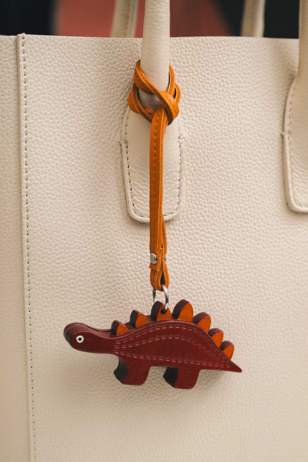 Dinosaur-shaped handmade leather bag charm attached to a beige handbag