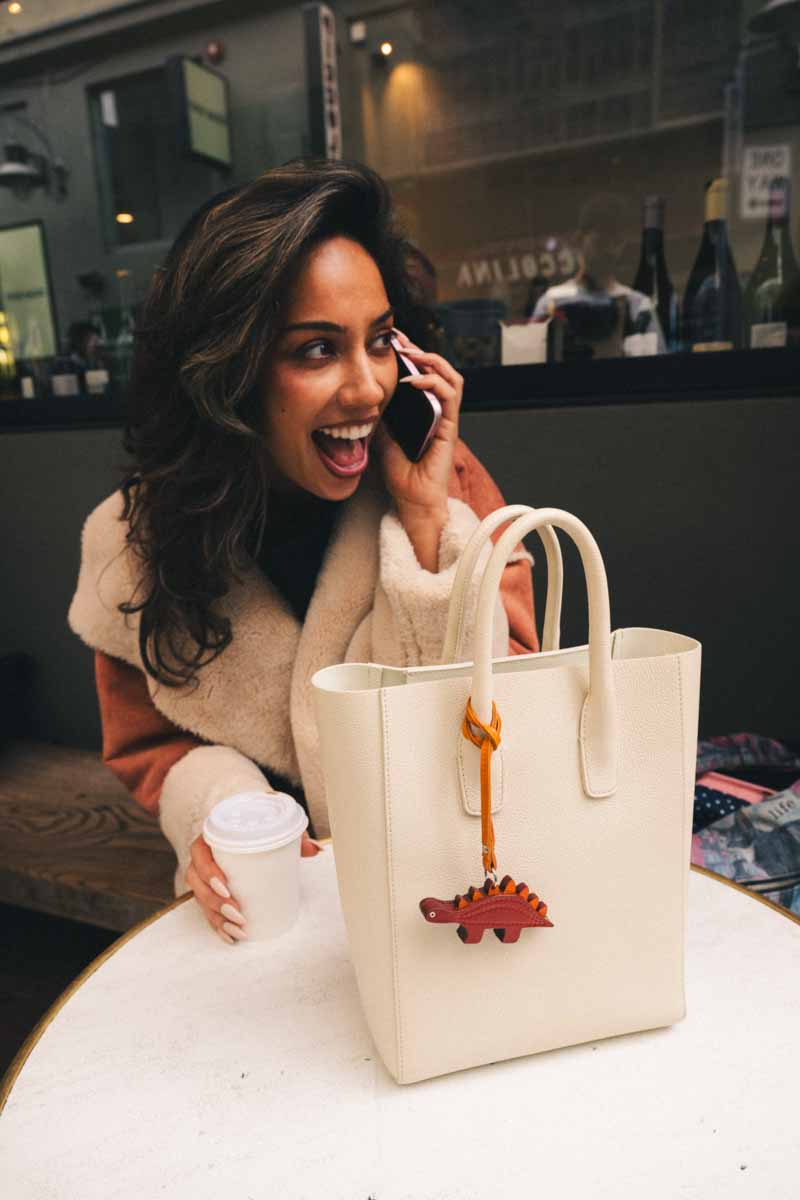 Woman talking on a phone with a beige handbag featuring a small brown dinosaur handmade leather bag charm on a table.
