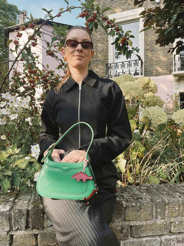 Person holding a green handbag with one pink and one green dinosaur bag charm, standing outdoors near a building and plants.