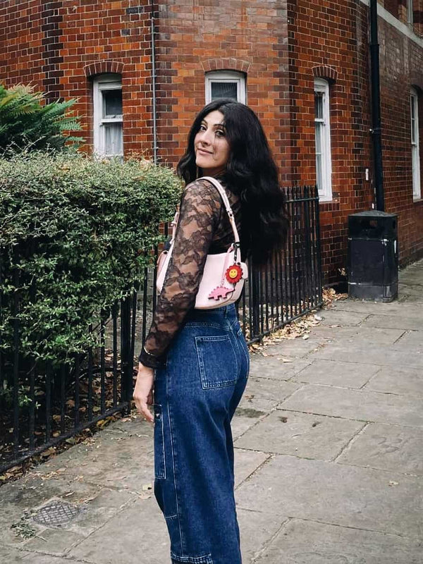 Woman with a pink handbag featuring smily flower and pink dinosaur bag charms standing on a sidewalk next to a brick building.