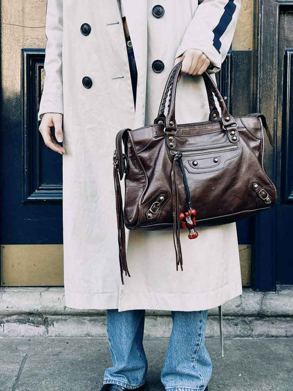 Person holding a brown leather handbag with a lobster bag charm, wearing a beige coat and blue jeans.