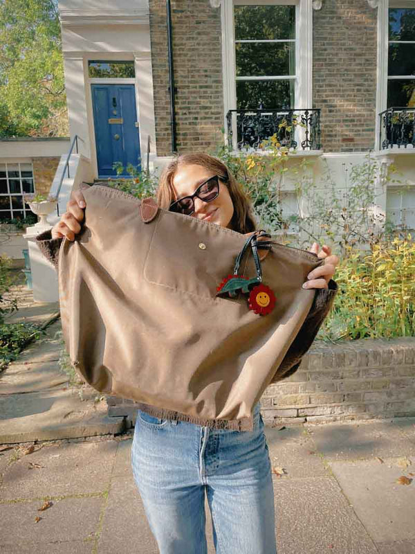Person holding a beige tote bag with one green dinosaur charm and one red flower charm in front of a house.