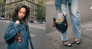 Woman wearing a denim jacket with a black handbag featuring dinosaur, lobster and floral handmade leather bag charm on a city street.